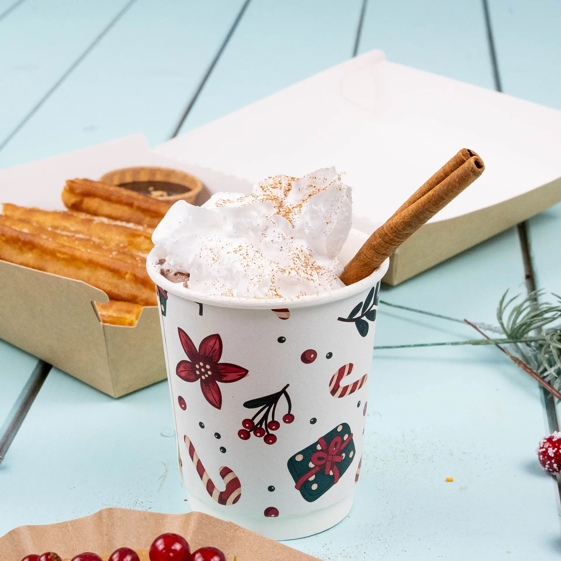 Doppelwandiger Becher mit Winterdesign, gefüllt mit Schlagsahne und einem Zimtstängel, neben Churros in einer Box.