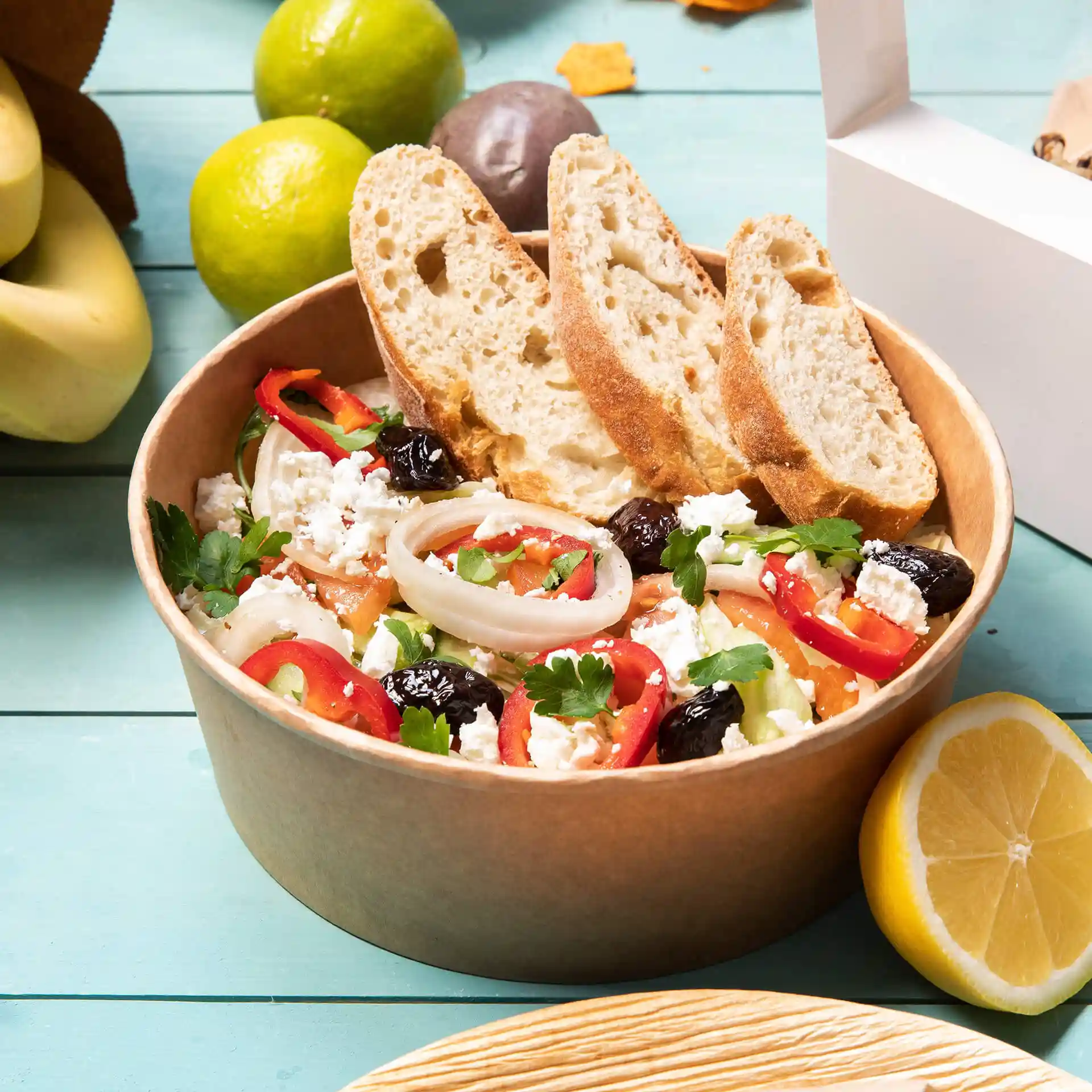Brown round paper bowl filled with salad, topped with sliced bread, surrounded by fruits and a white takeout box.