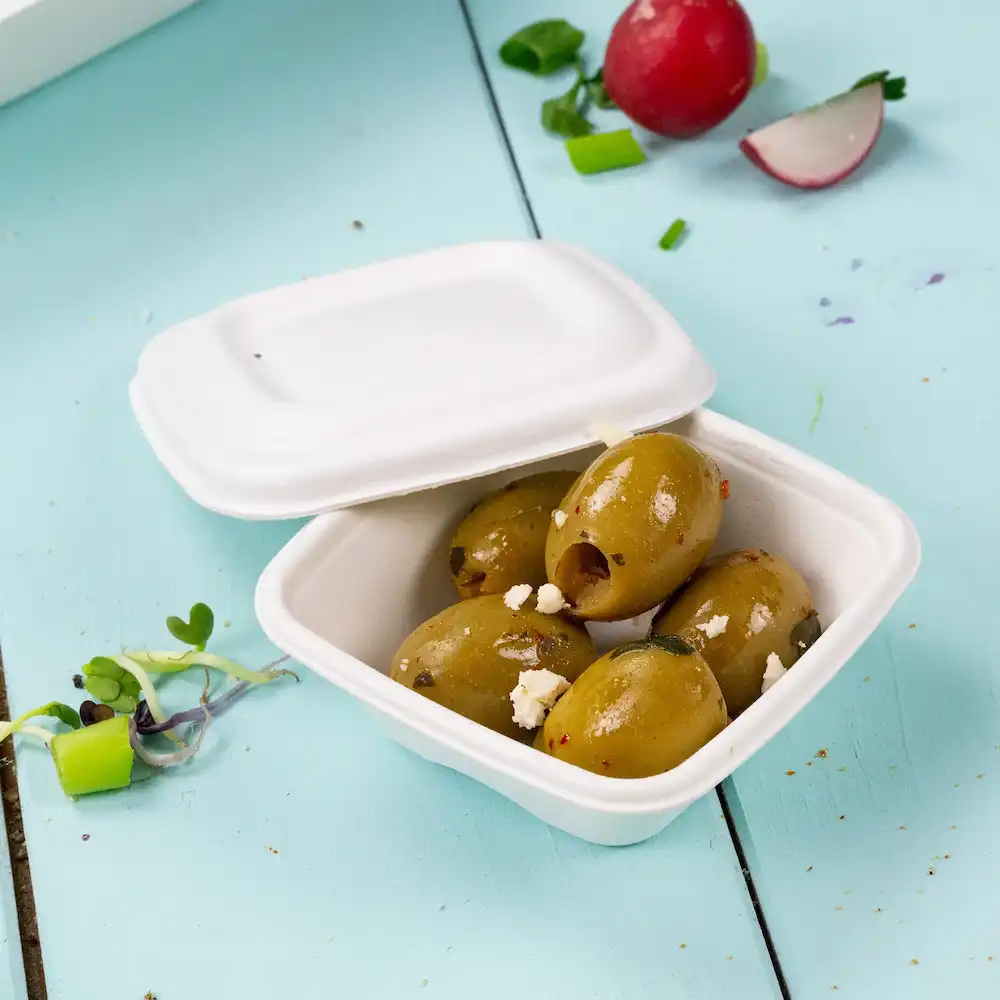 Container holding several green olives with toppings, placed on a light blue background, with a radish and herbs nearby.