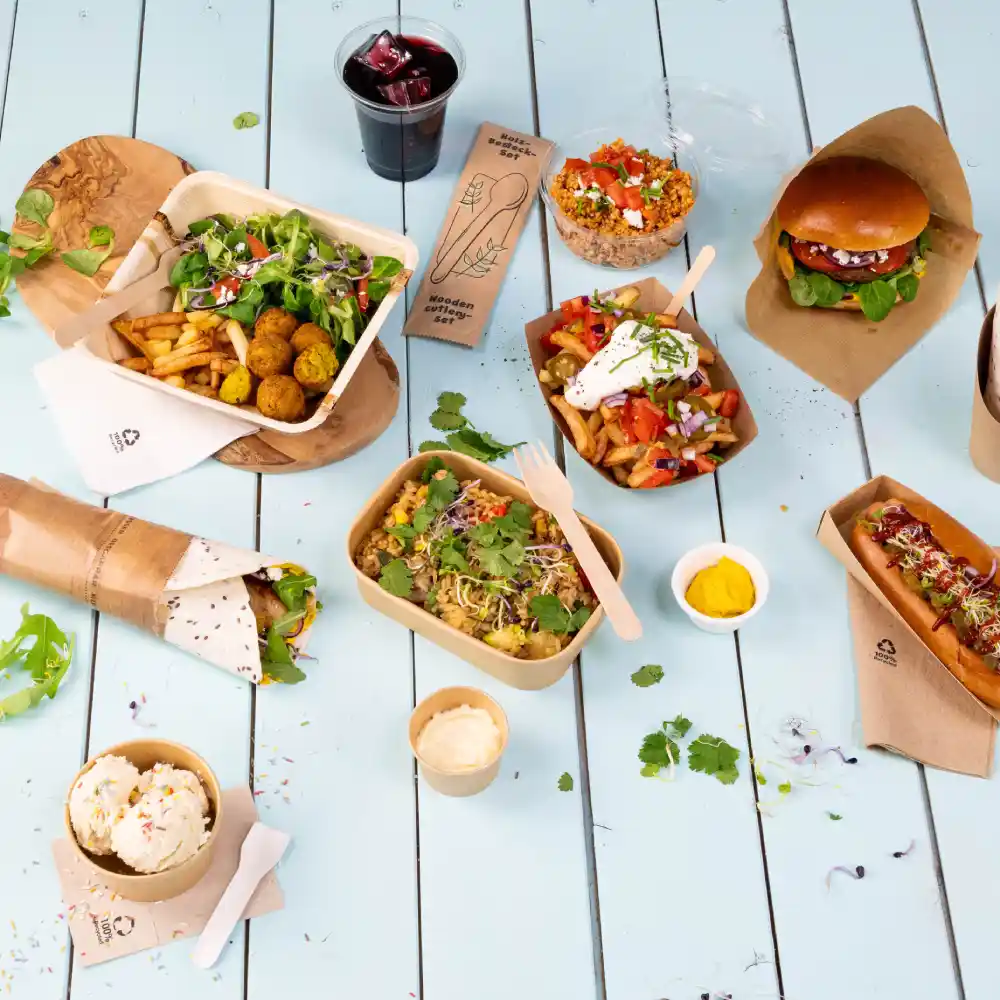 Anordnung verschiedener Catering-Gerichte auf einem hellblauen Tisch, einschließlich Salaten, Burgern, Hot Dogs und Desserts. Ein Getränk ist ebenfalls enthalten.