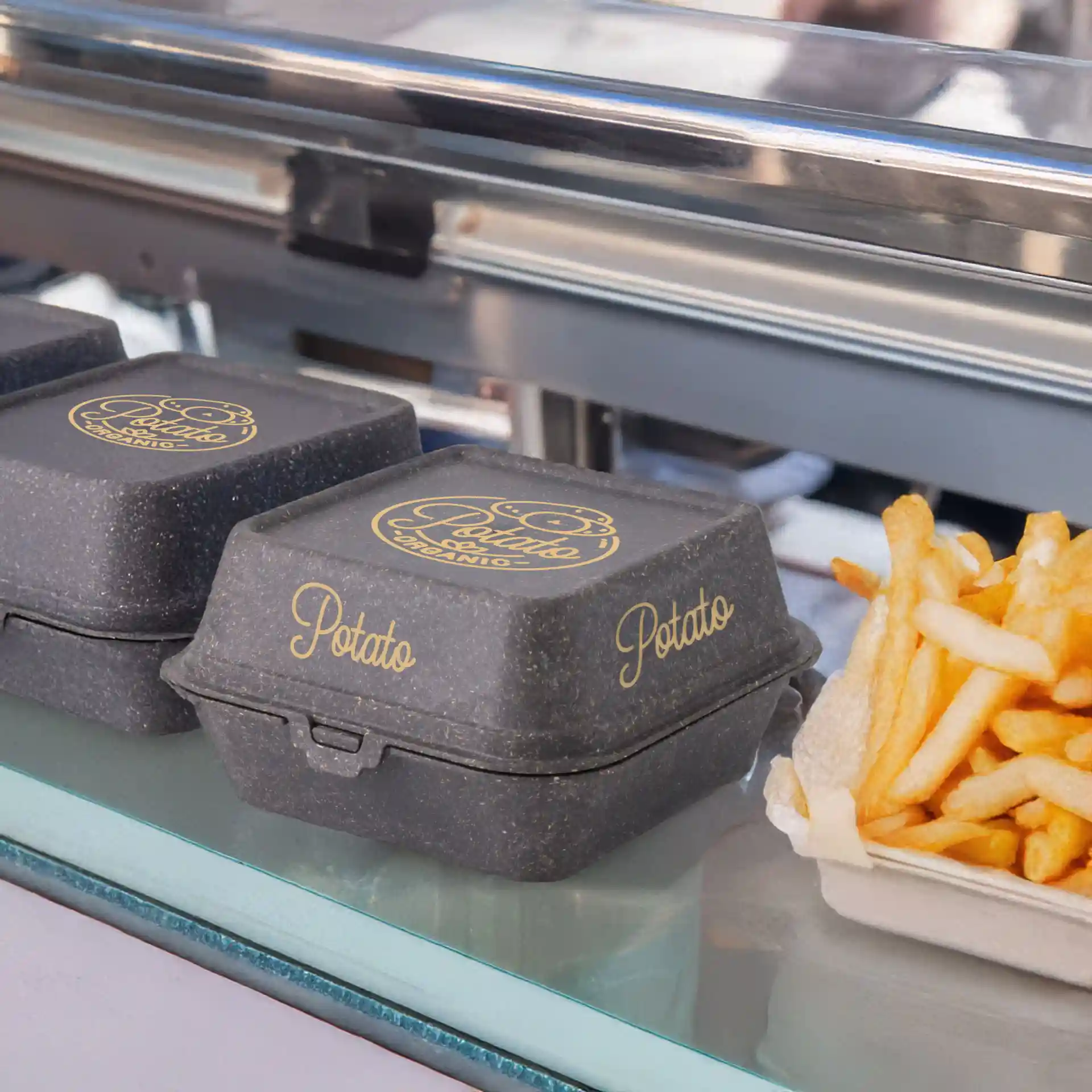 Grau bedruckte wiederverwendbare Burgerboxen in rechteckiger Form, neben Pommes auf einer Glasoberfläche präsentiert.