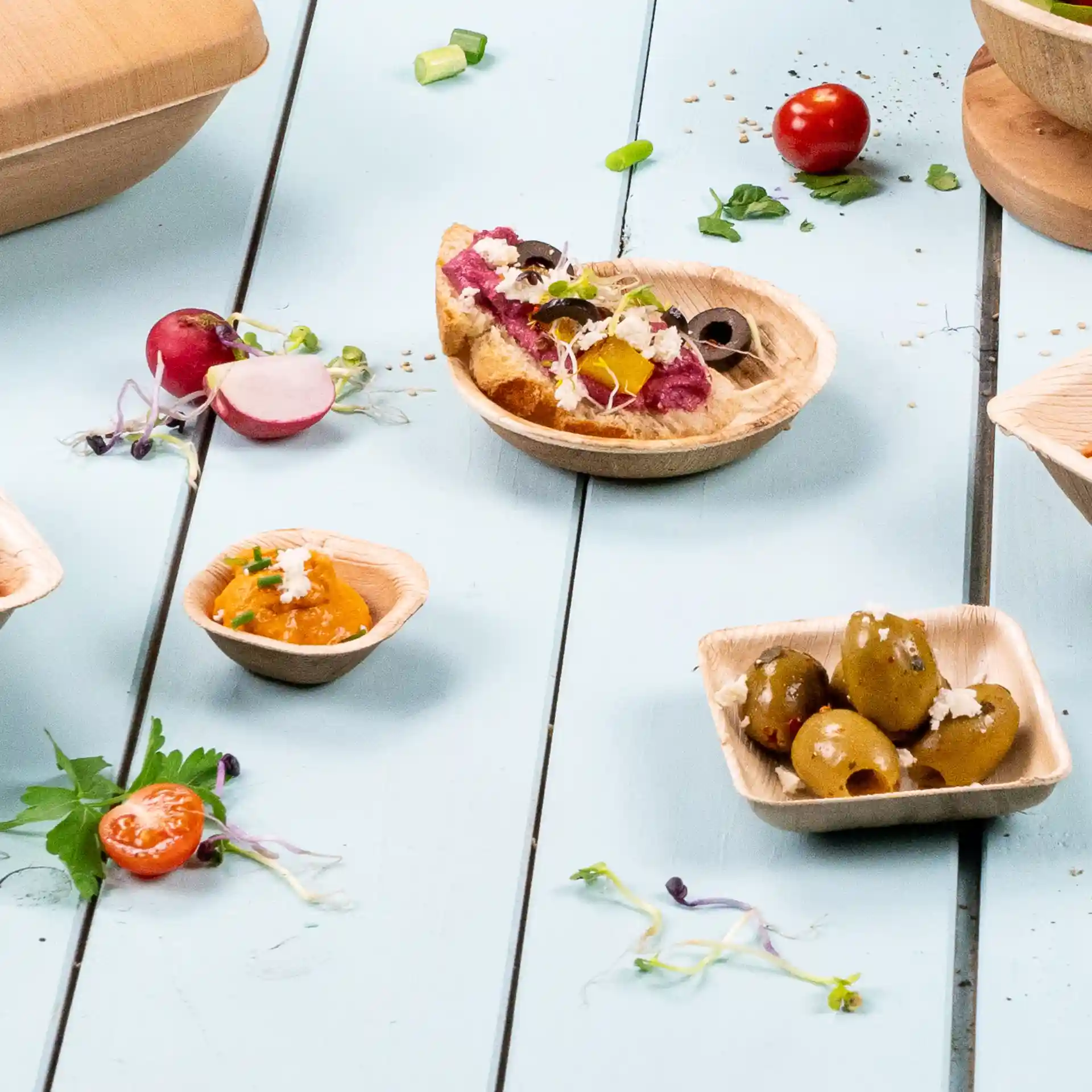 Image of round palm leaf bowls with various food items, including olives, dips, and garnishes on a blue wooden surface.