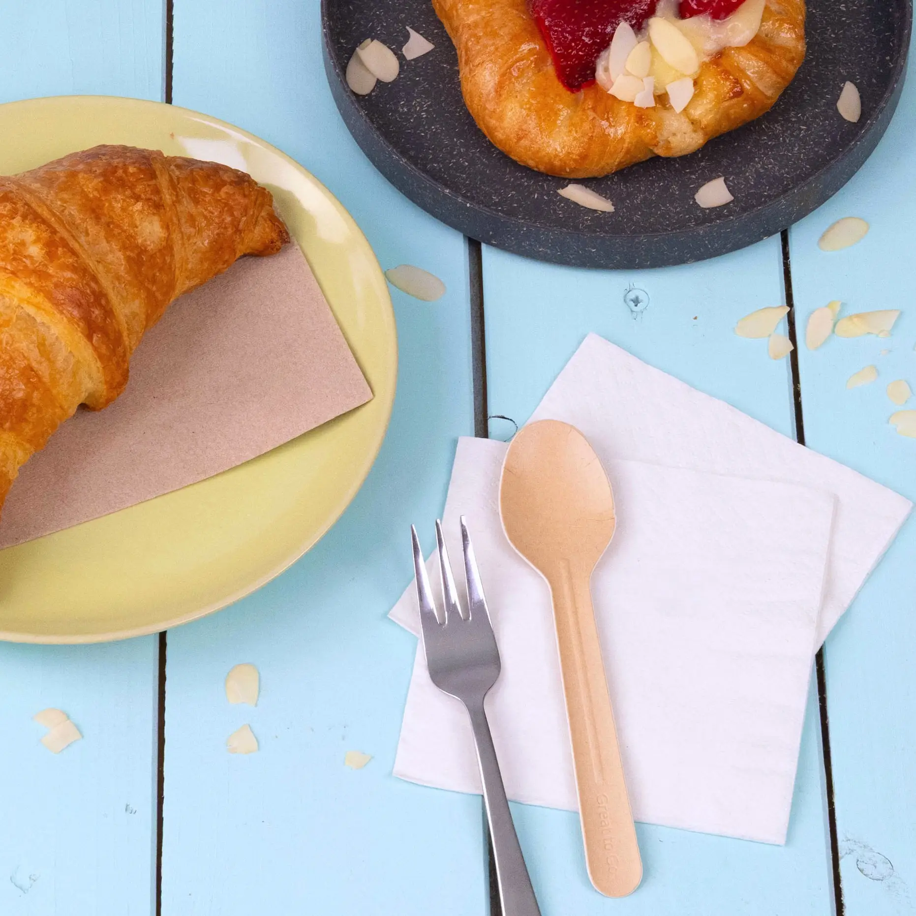 Bild eines Croissants auf einem gelben Teller, einem Gebäck auf einem dunklen Teller, einem Papierspoon, einer Gabel und Servietten auf einem blauen Holztisch.