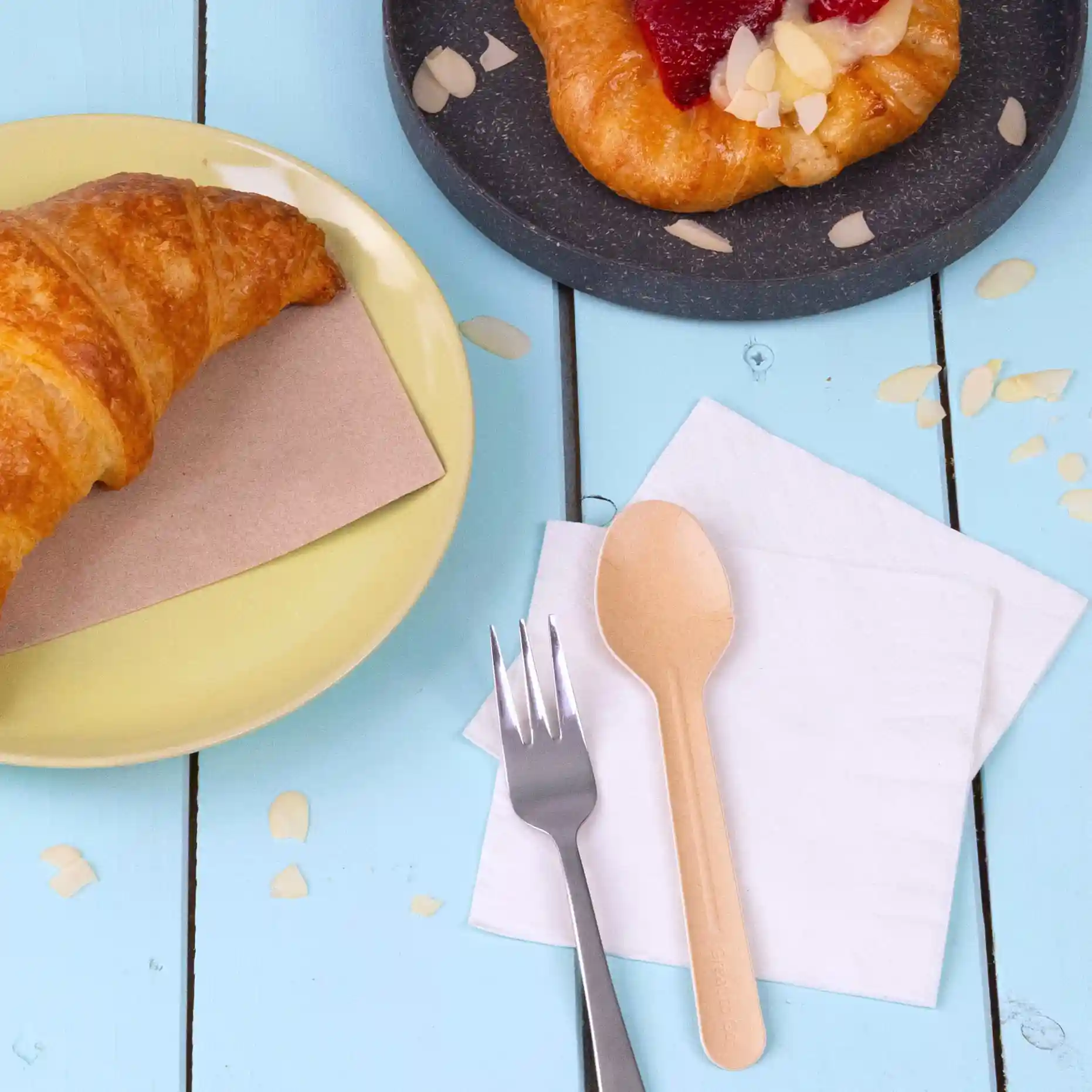 Image of a croissant on a yellow plate, a pastry on a dark plate, a paper spoon, a fork, and napkins on a blue wooden table.
