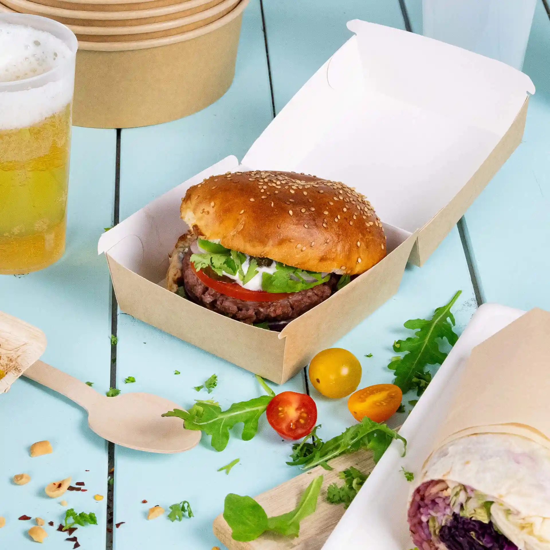 Braune Pappbox für Burger mit einem Sesambrötchen, Salat, Tomate und Sauce auf einer blauen Oberfläche.