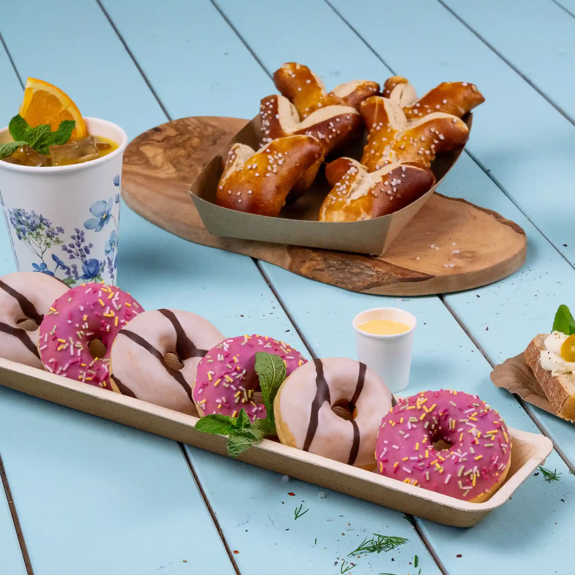 Rechteckiges Palmblatt-Tablett mit mehreren Donuts, einem Getränk und Brezeln auf einer blauen Oberfläche.