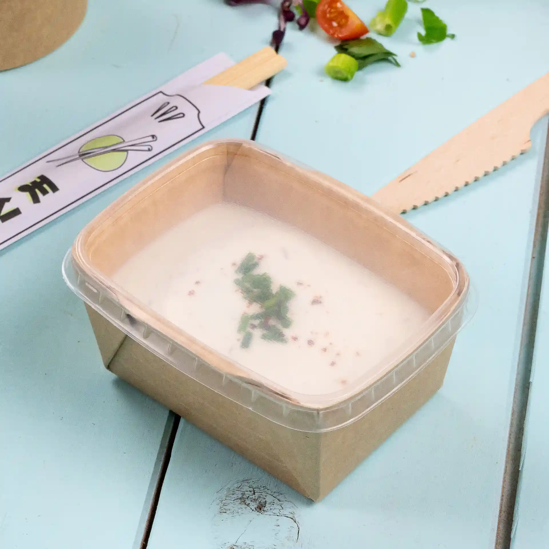 Transparenter rechteckiger Becher mit klarem Deckel, gefüllt mit cremiger Suppe und grünen Kräutern.