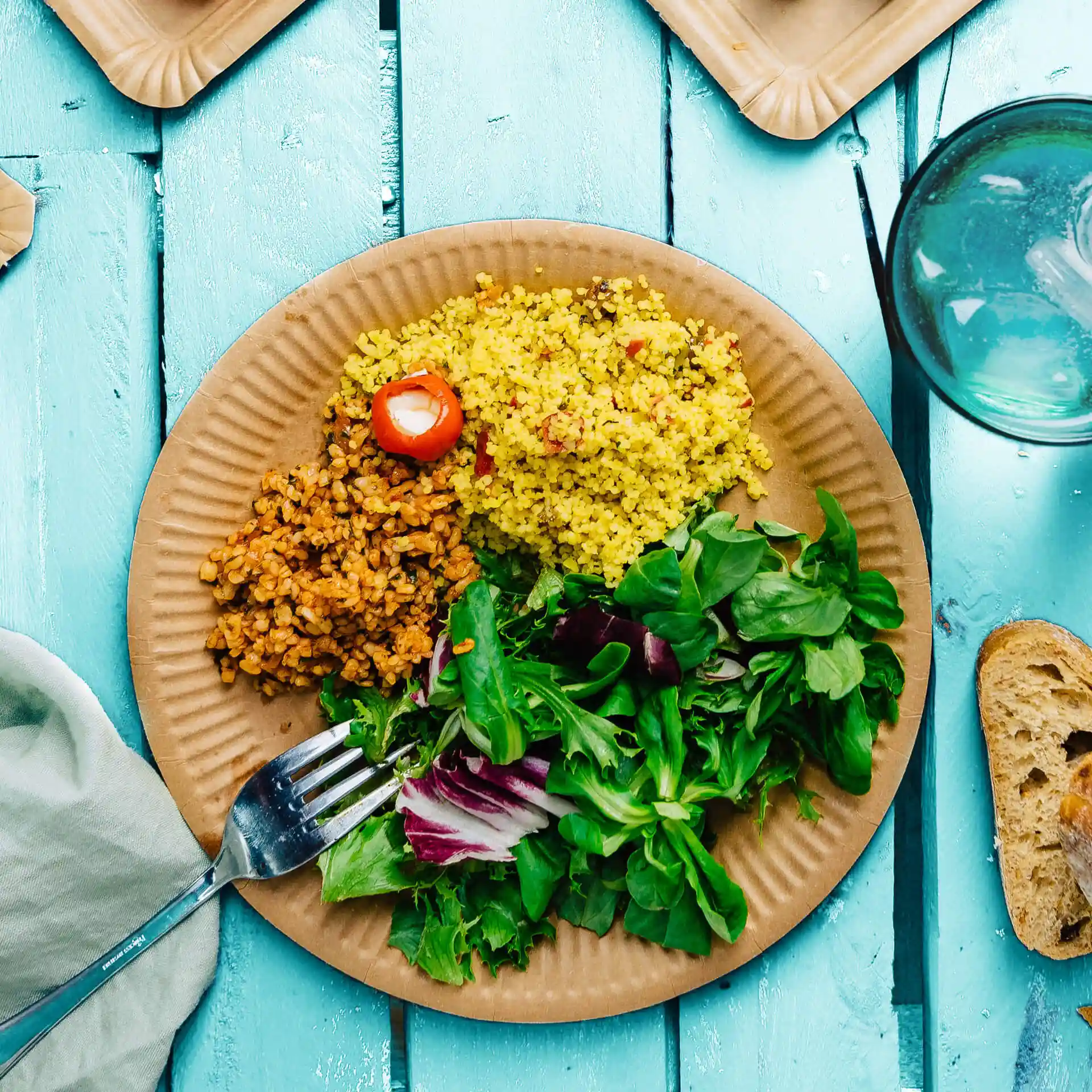 Plate with yellow couscous, brown grains, and mixed greens, served on a round brown paper plate.