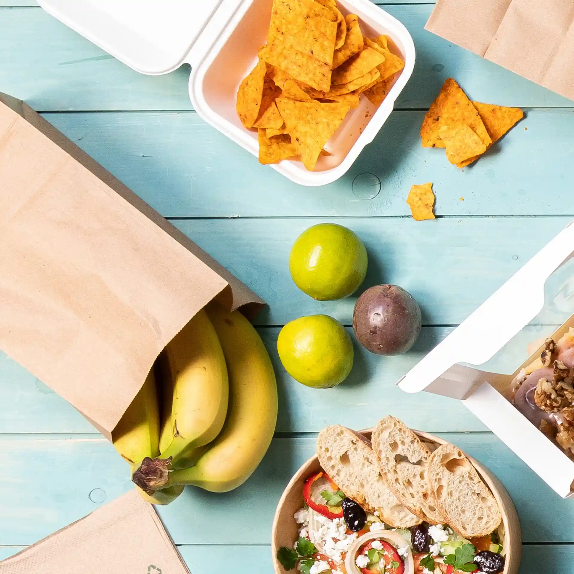 Bild einer braunen Kraftpapiertüte mit Bananen, einer Schüssel Salat, Limetten, einer Passionsfrucht und einem Behälter mit Chips auf einer blauen Holzoberfläche.