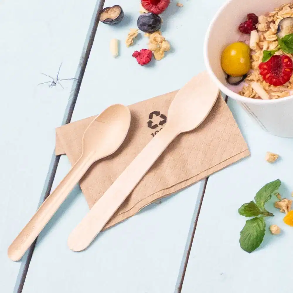 Two wooden spoons placed on a brown napkin beside a cup of granola topped with fruits and mint leaves.