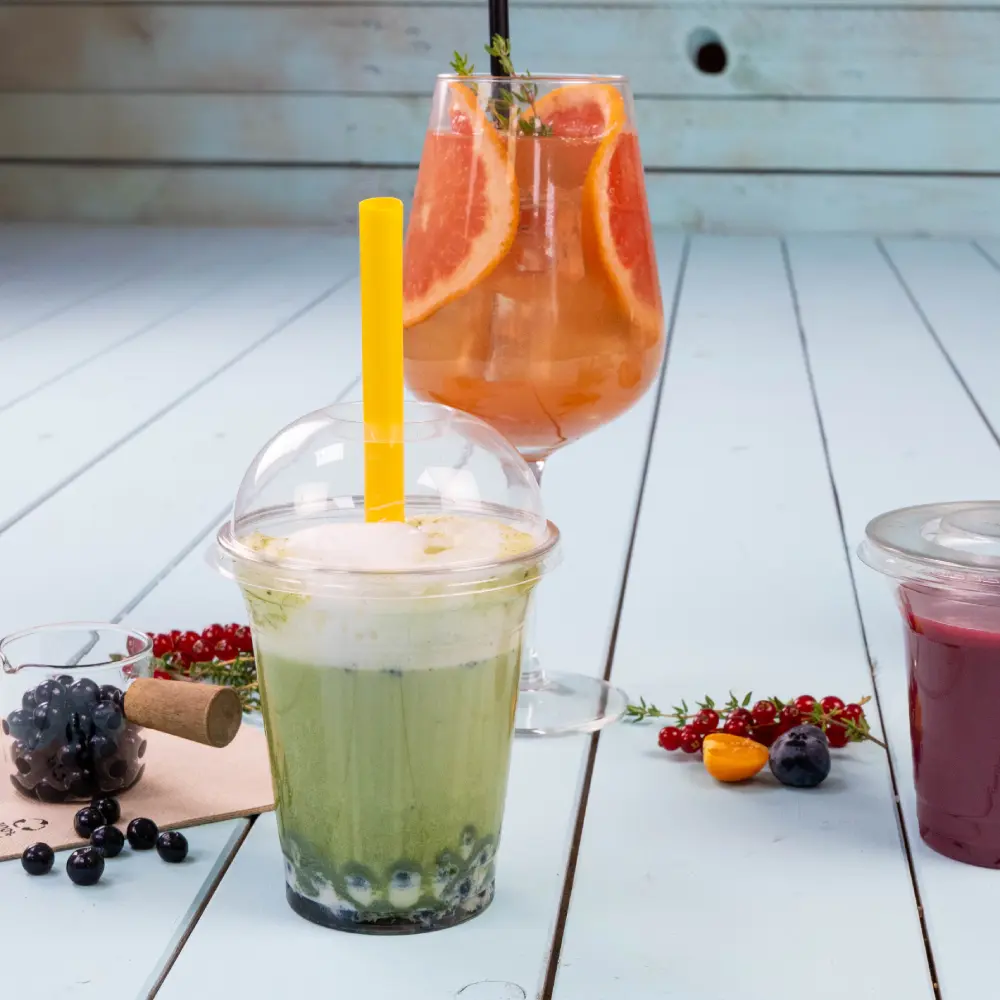 Ein grünes Getränk mit einem gelben Strohhalm in einem klaren Becher, mit blauen Tapiokaperlen am Boden; ein bunter Cocktail mit Grapefruitscheiben in einem Glas dahinter.