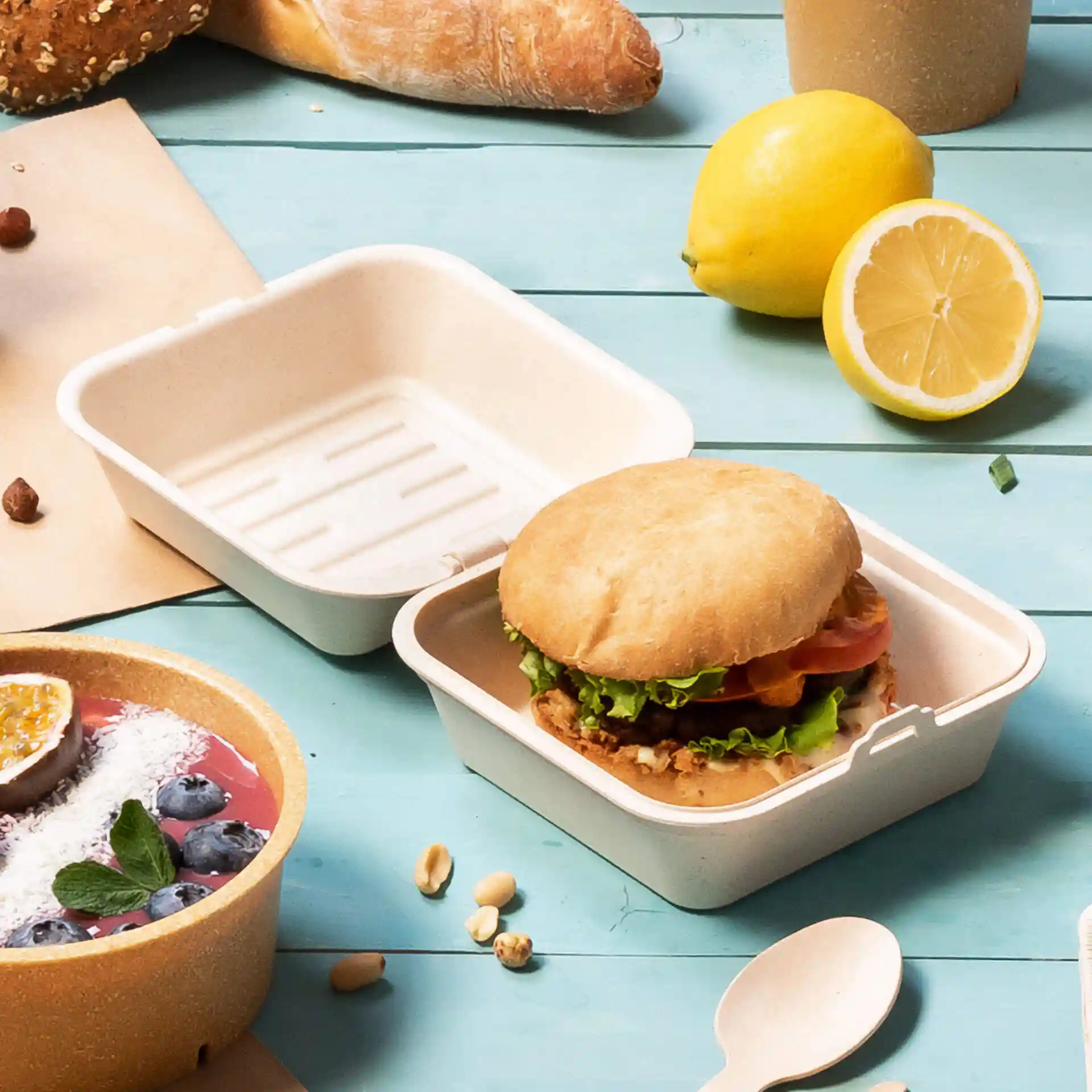 Wiederverwendbare Burgerbox in cremeweiß mit einem Burger darin, umgeben von Lebensmitteln auf einer blauen Oberfläche.