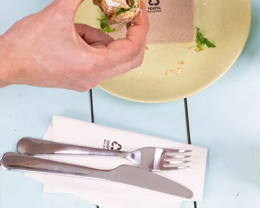 Hand holding a sandwich above a pale yellow plate, with a knife and fork on a napkin beside it.