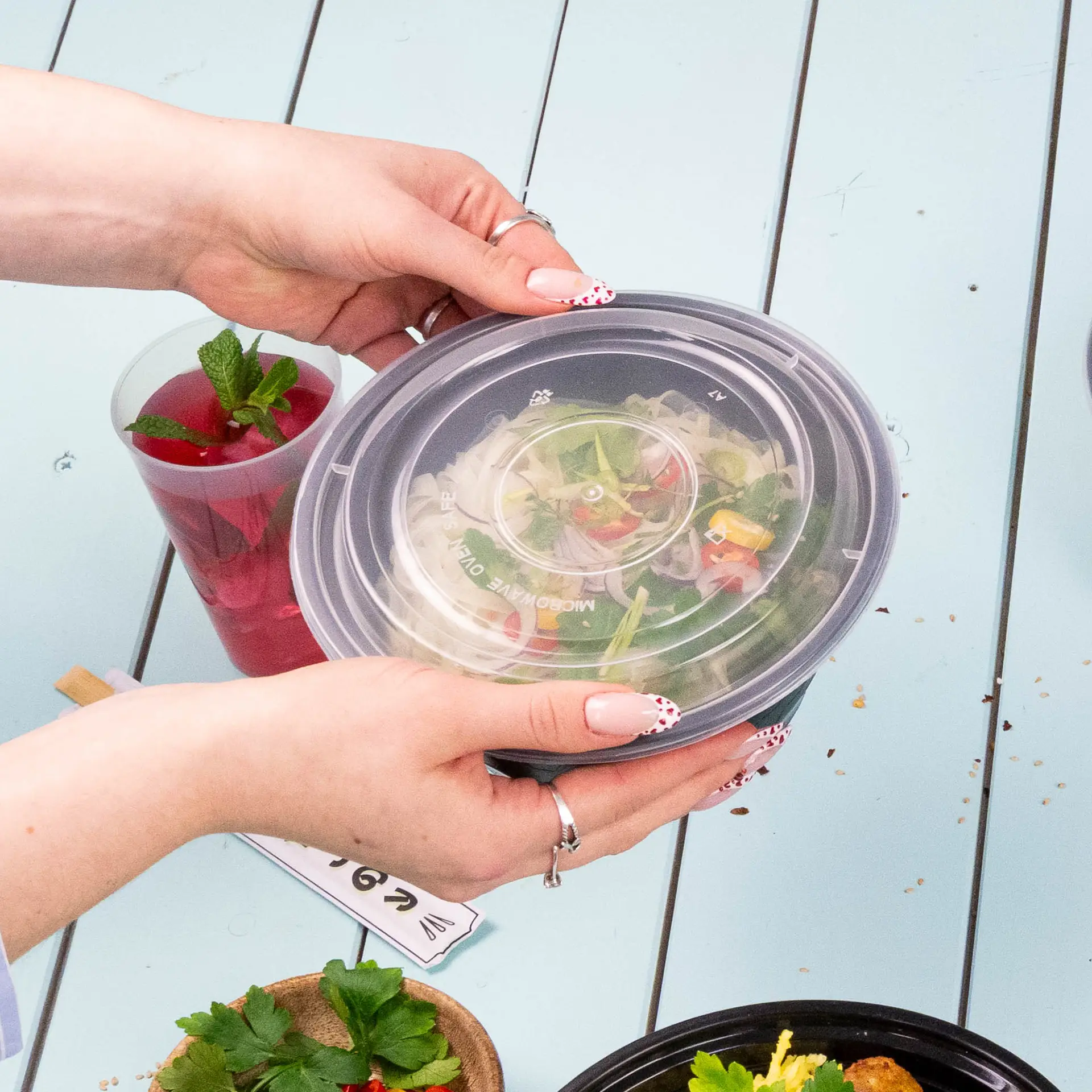 One transparent reusable lid for food container held by hands; includes a colorful salad and a drink in the background.