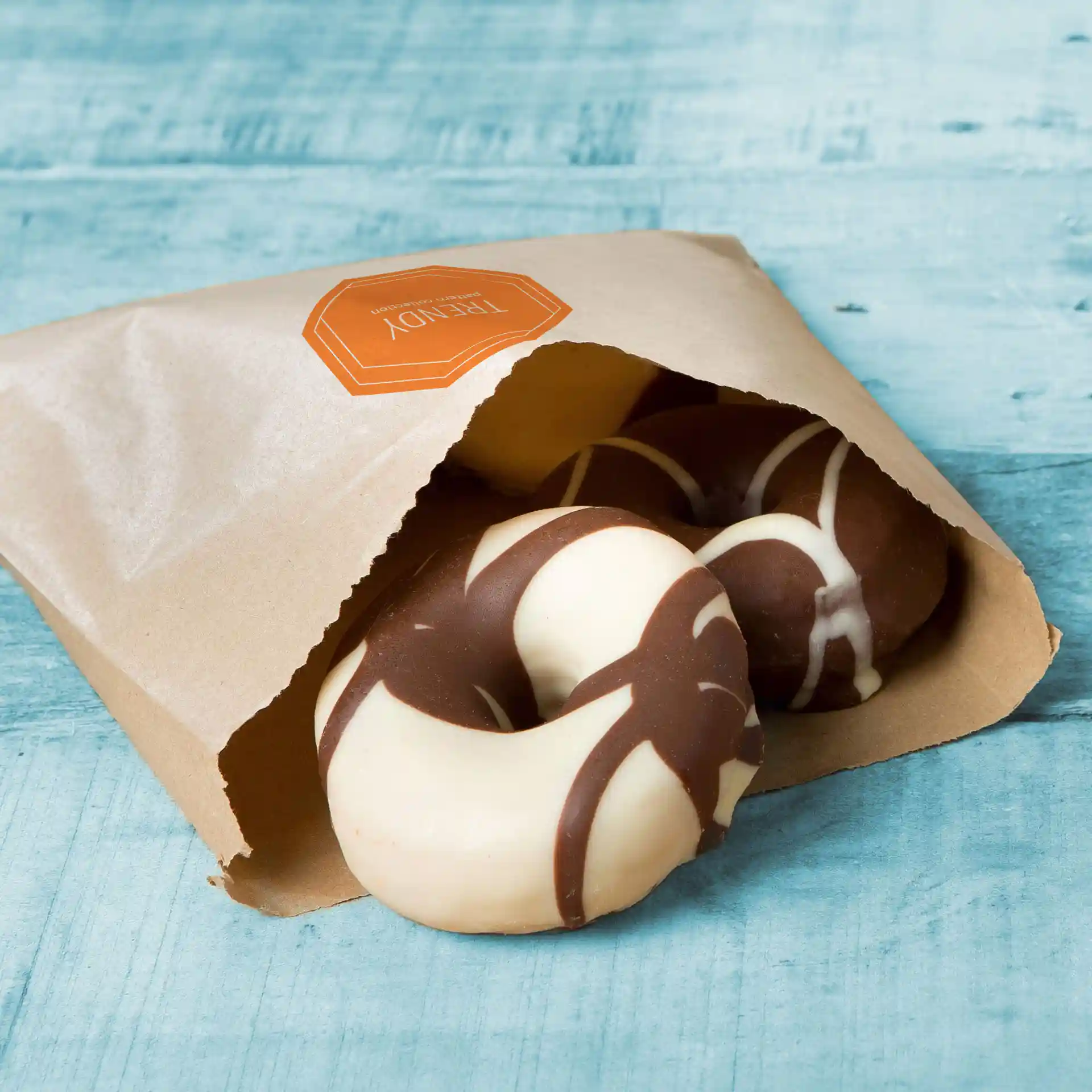 Braune flache Papiertüte mit teilweise sichtbaren schokoladenüberzogenen Donuts, auf einer blauen Holzoberfläche.