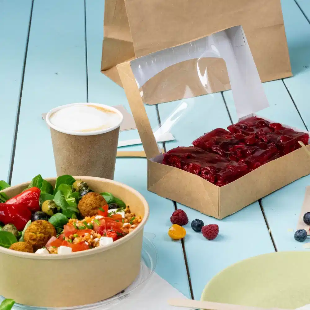 Ein Takeaway-Mahlzeiten-Setup mit einem Salat in einer Schale, einem Becher und einem Dessert in einer transparenten Box auf einem Holztisch.