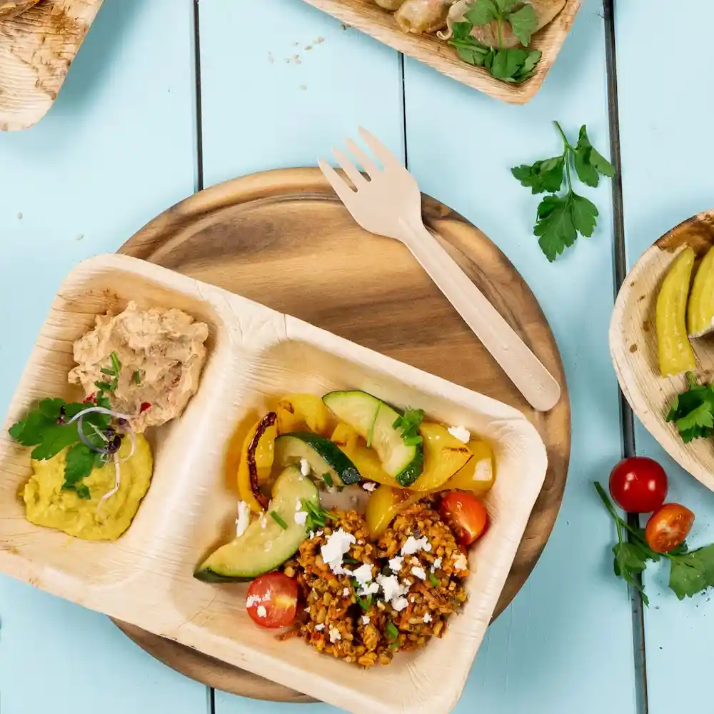 Disposable wooden fork next to a plate containing hummus, vegetables, and a grain dish on a wooden surface.