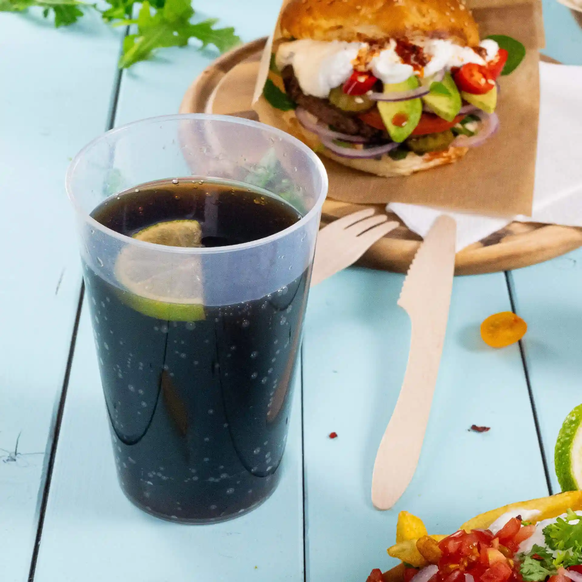 Transparent reusable cup filled with dark beverage and a lime slice, placed beside a burger and wooden utensils.