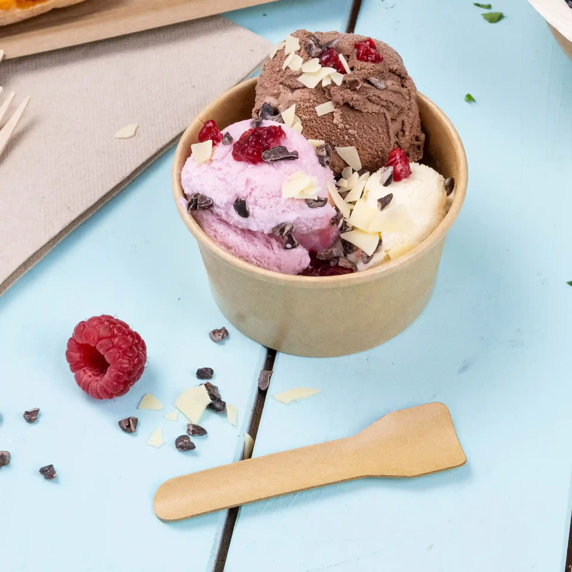 Becher mit drei Kugeln Eis in Pink, Braun und Weiß, garniert mit Schokoladenstückchen und Himbeeren, mit einem Holzlöffel.