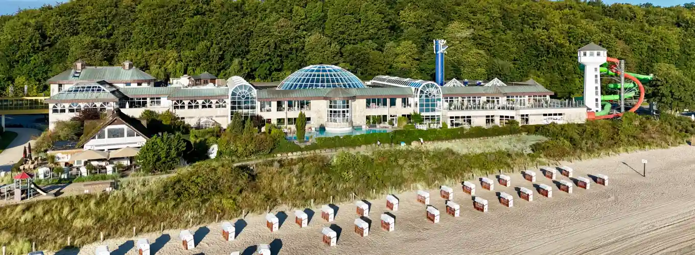 Aerial view of Ostsee Therme building near a beach, featuring a glass dome and water slides, with beach huts in the foreground.