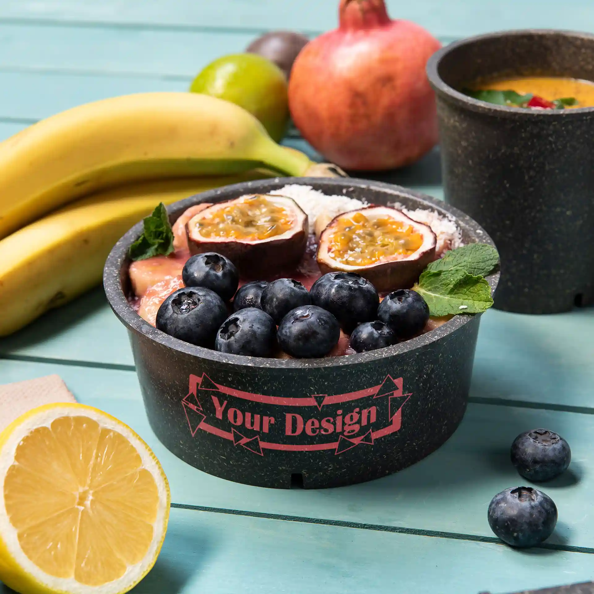 Runde graue wiederverwendbare Schüssel mit Obsttoppings, darunter Heidelbeeren und Passionsfrucht, auf einer türkisfarbenen Oberfläche.