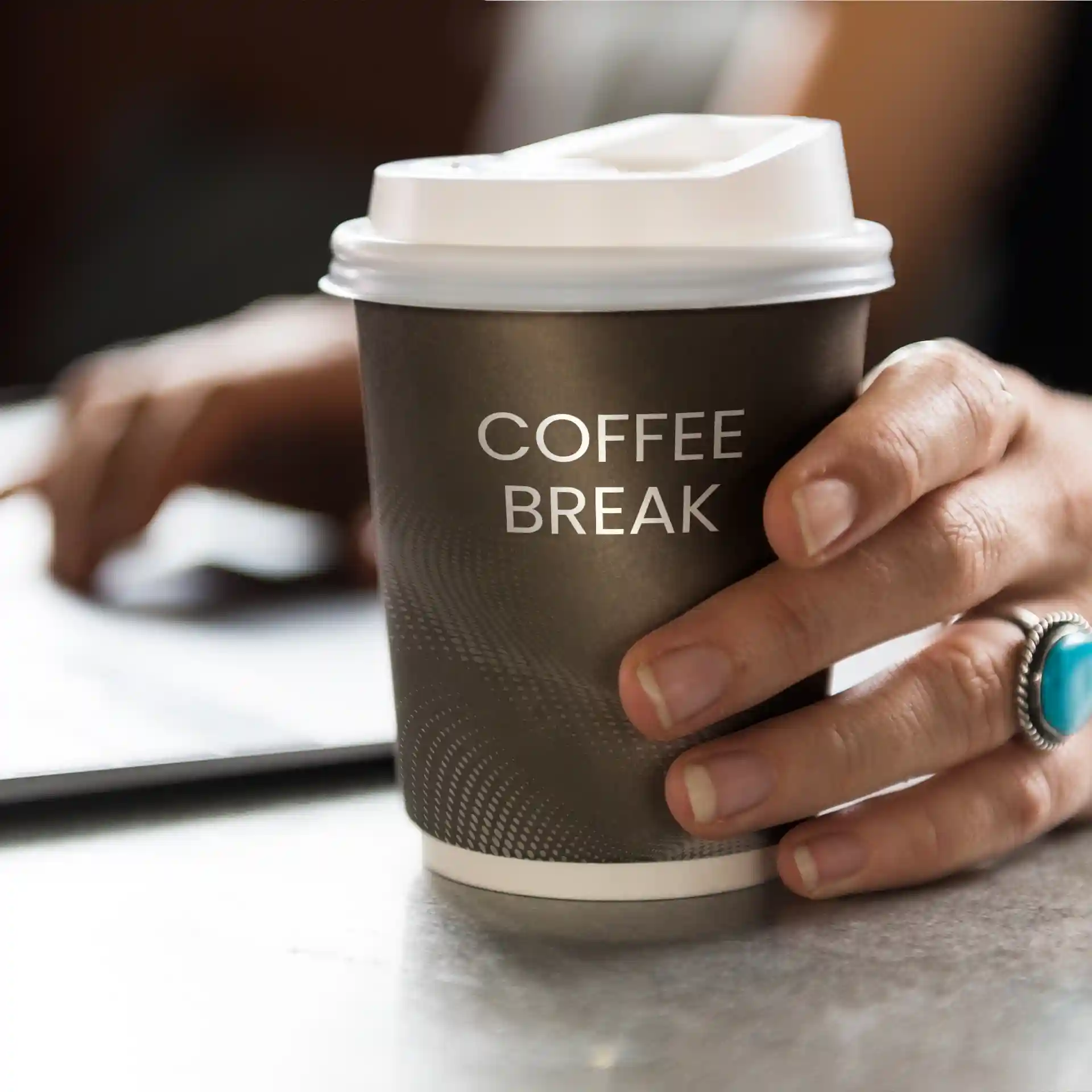 Brown paper coffee cup with a white lid, held in a hand, featuring the text "COFFEE BREAK".