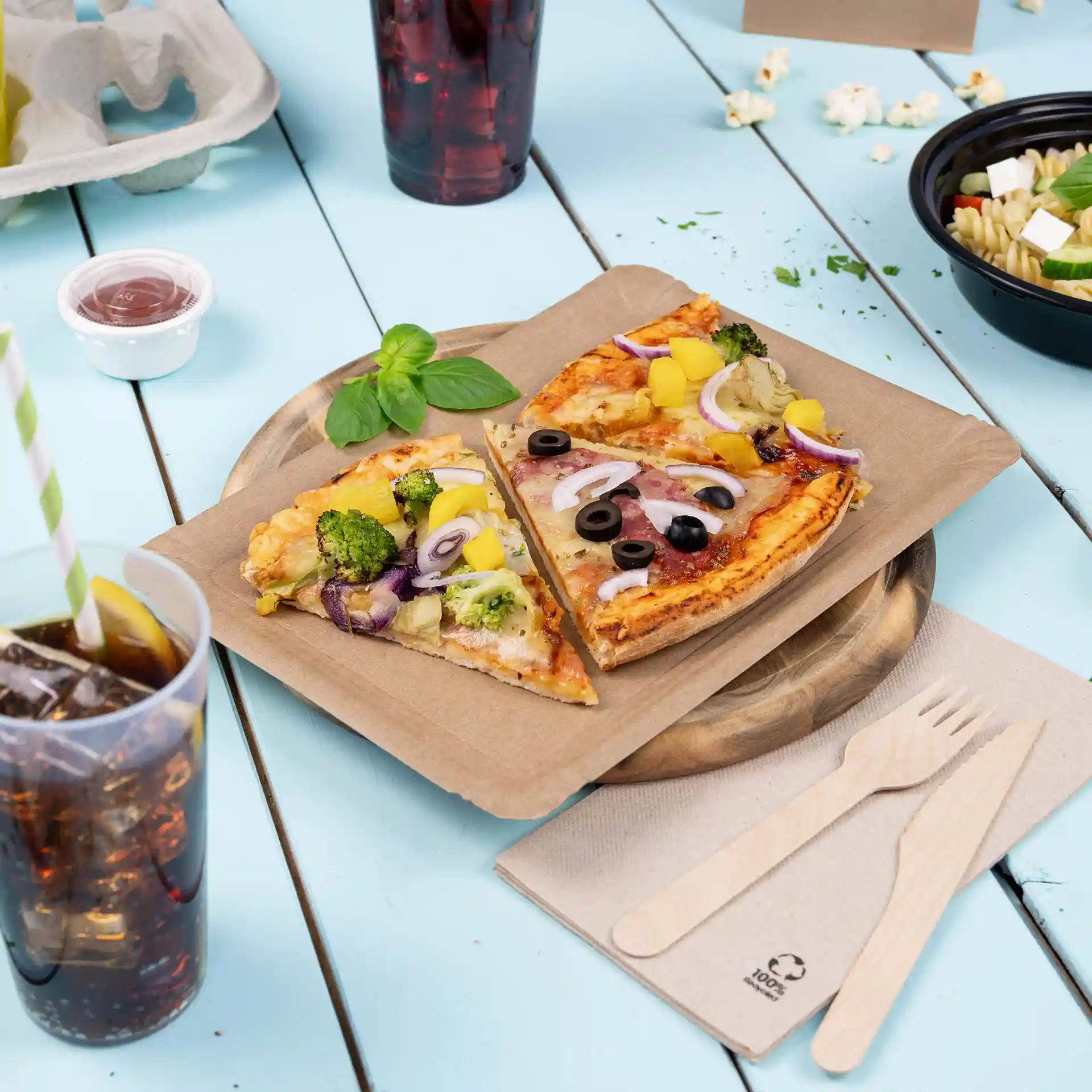 Bild von zwei Pizzastücken mit Belag auf einem braunen rechteckigen Pappteller, begleitet von einem Getränk und Besteck auf einem blauen Tisch.
