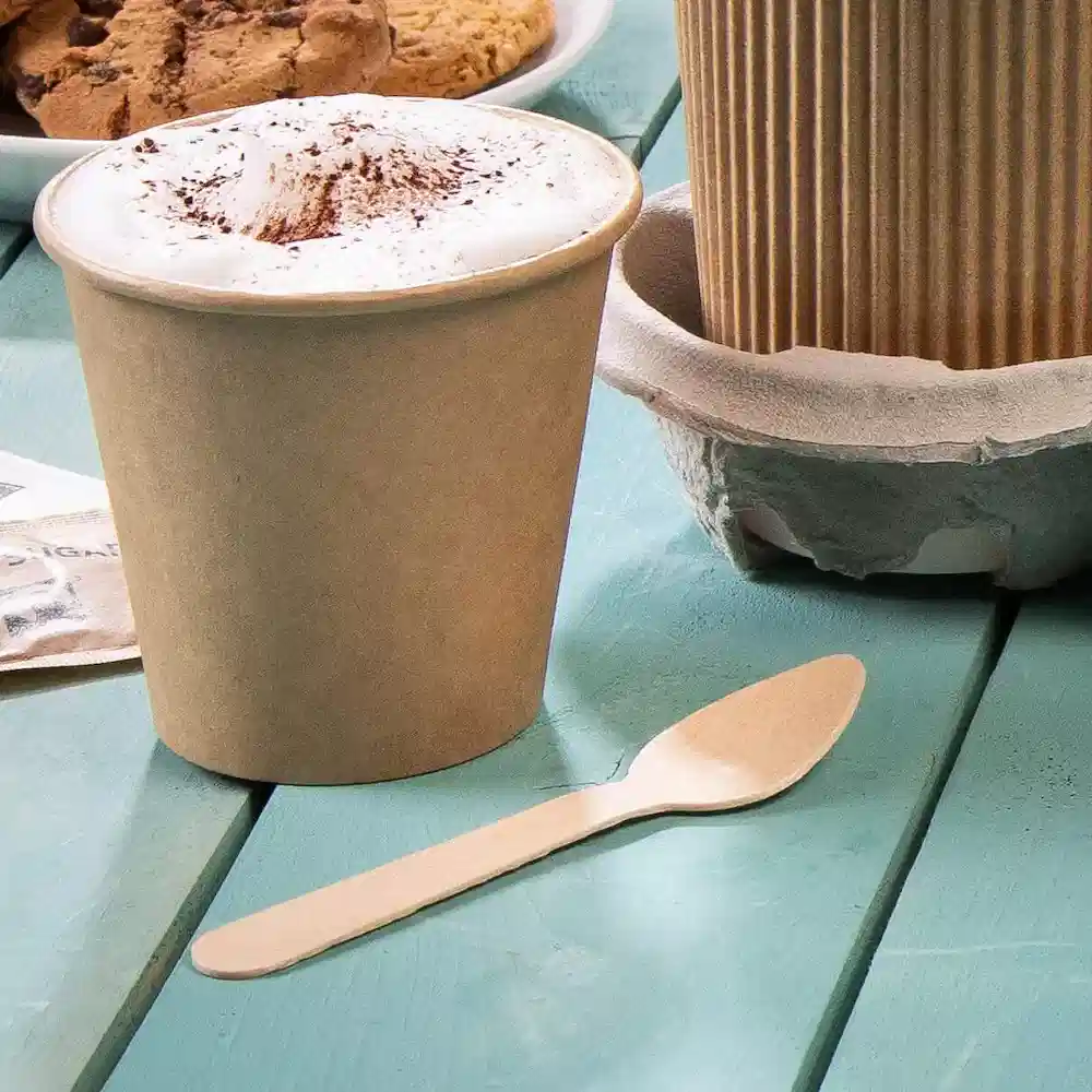 Brauner Einwegbecher mit Schaumdecke und einem Holzlöffel auf einem türkisfarbenen Tisch, im Hintergrund Kekse.