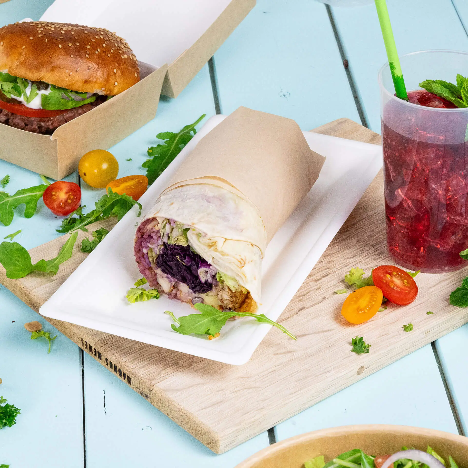 Dish featuring a wrap on a rectangular sugarcane plate, with a burger, drink, and various vegetables in the background.