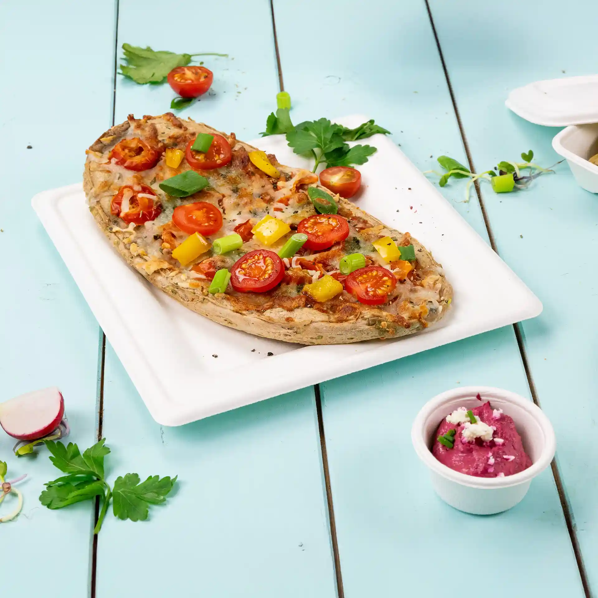 Quadratischer weißer Becher aus Zuckerrohr mit Pizza, belegt mit Tomaten, Paprika und Frühlingszwiebeln, neben einem kleinen Becher Dip.