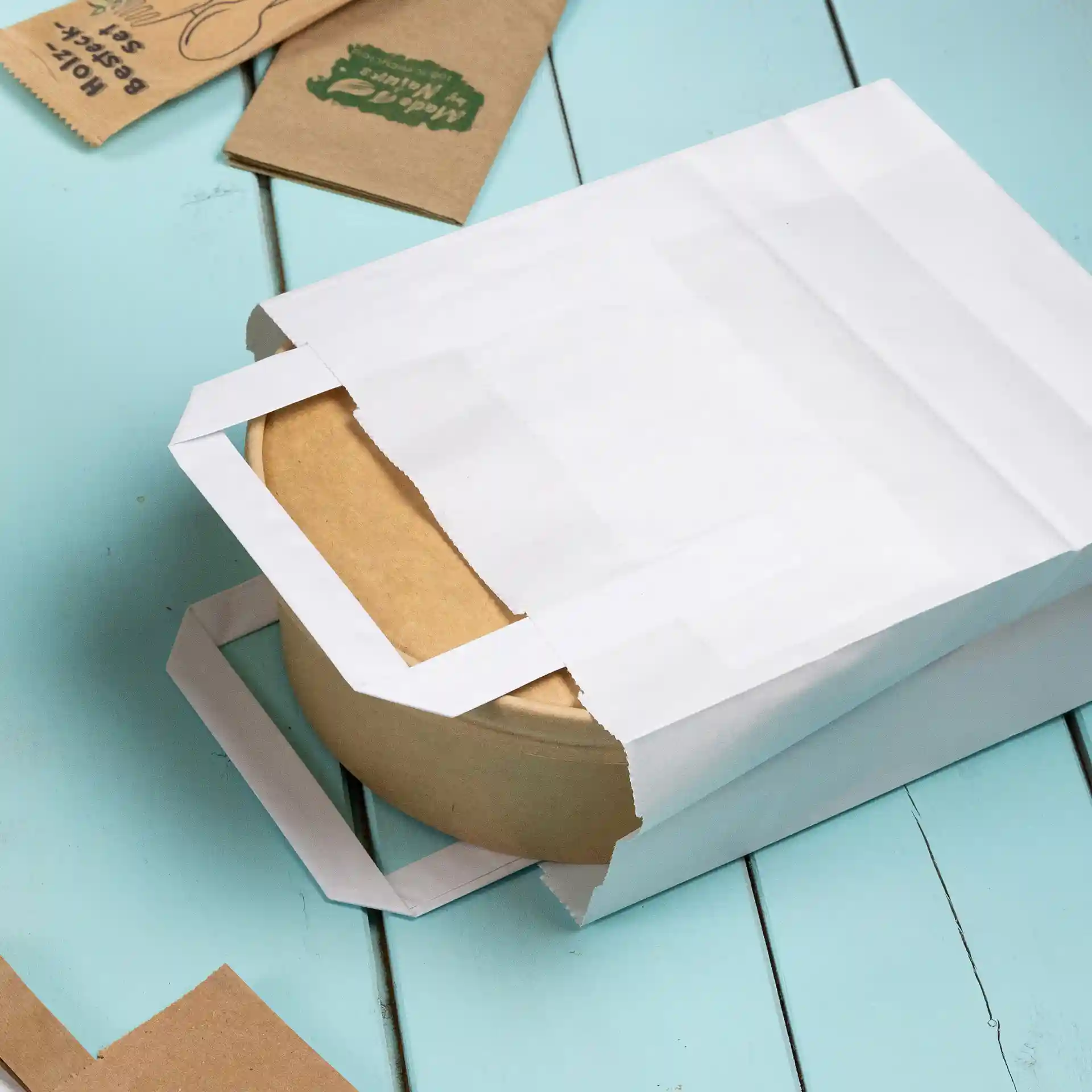 Weißer Kraftpapierbeutel teilweise geöffnet, mit einem runden braunen Behälter darin, auf einer hellblauen Oberfläche.