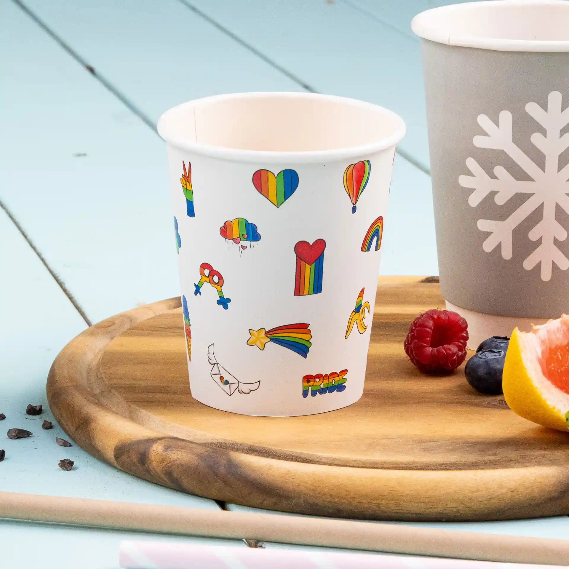 Weißer Becher mit bunten Druckdesigns, darunter Herzen, Regenbögen und Wolken, auf einem Holztablett.