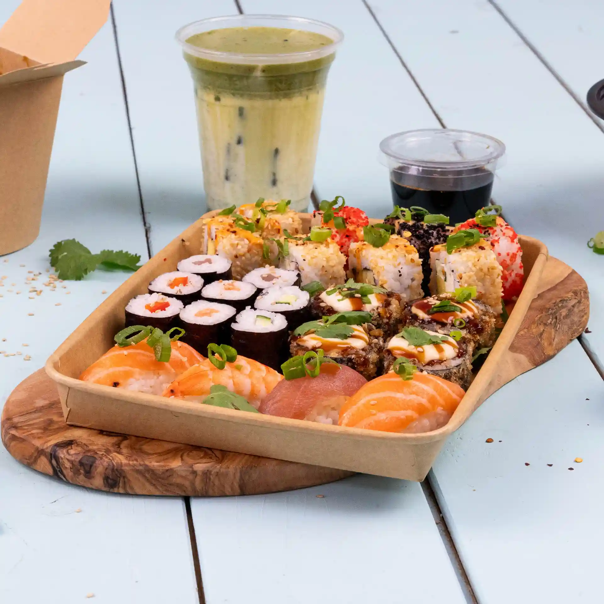 Tablett mit verschiedenen Sushi-Rollen, darunter Garnelen und Lachs, mit einem Getränk und Dip-Sauce auf einem Holzbrett.