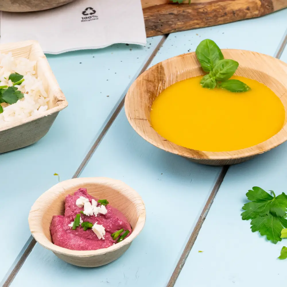 Bambusschüssel gefüllt mit gelber Suppe, garniert mit einem Basilikumblatt; kleinere Schüssel mit rosafarbener Dipsauce, garniert mit Kräutern; Reis mit Kräutern in einem Behälter daneben.