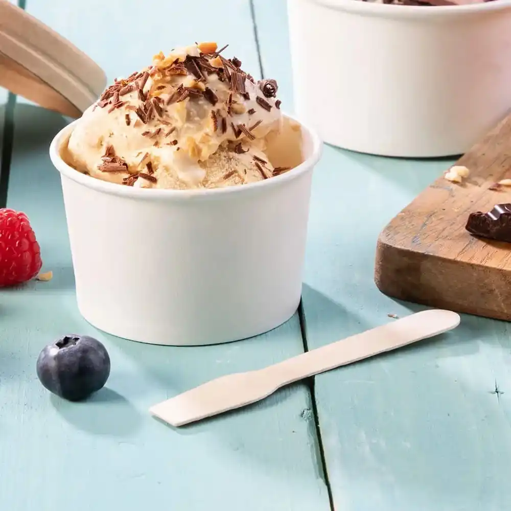 Weiße Einweg-Löffel neben einem Becher Eiscreme, garniert mit Schokolade und Nüssen, mit Beeren auf einer blauen Holzoberfläche.