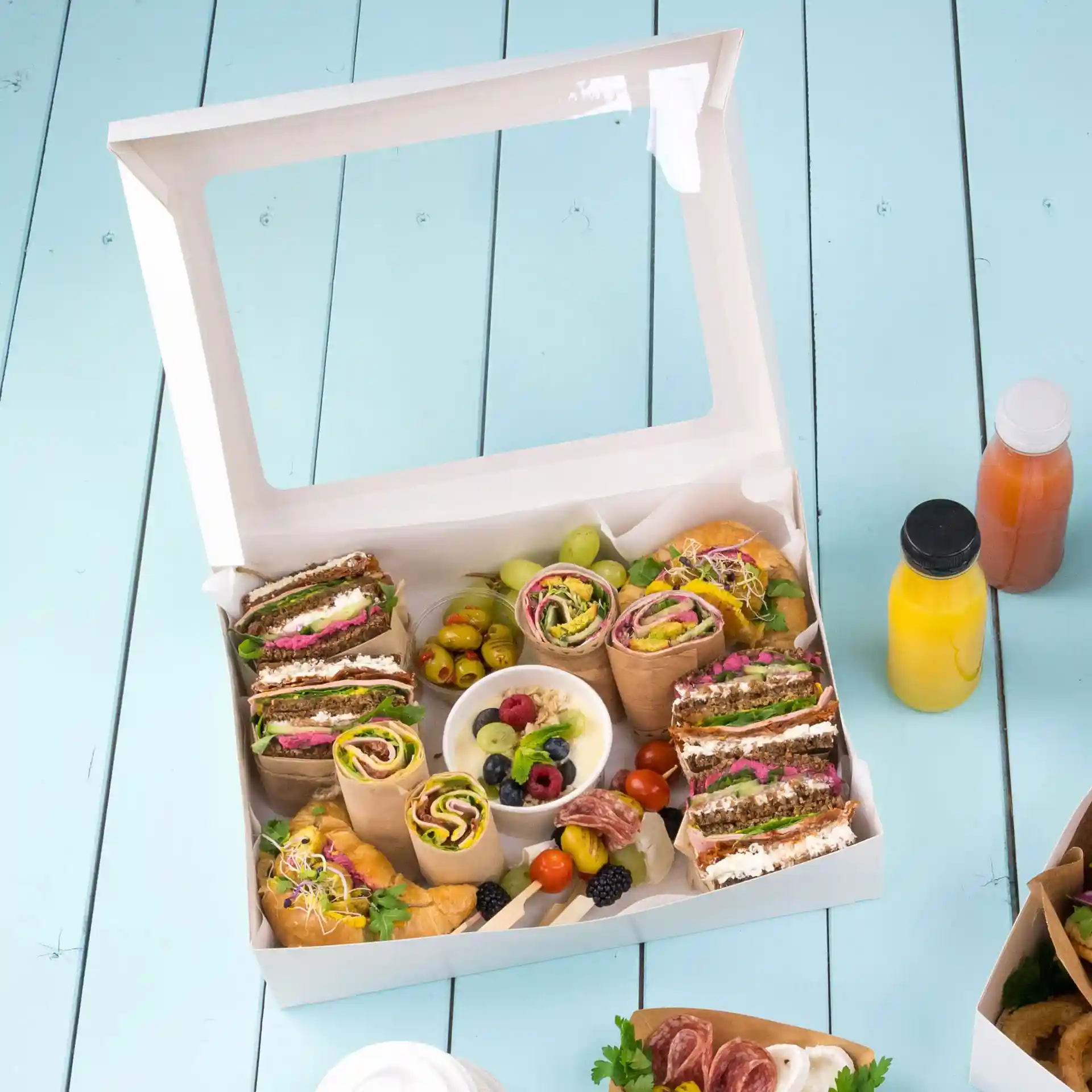 Box mit verschiedenen Wraps, Sandwiches, Früchten und einer kleinen Schüssel mit gemischten Beeren auf einer hellblauen Oberfläche.