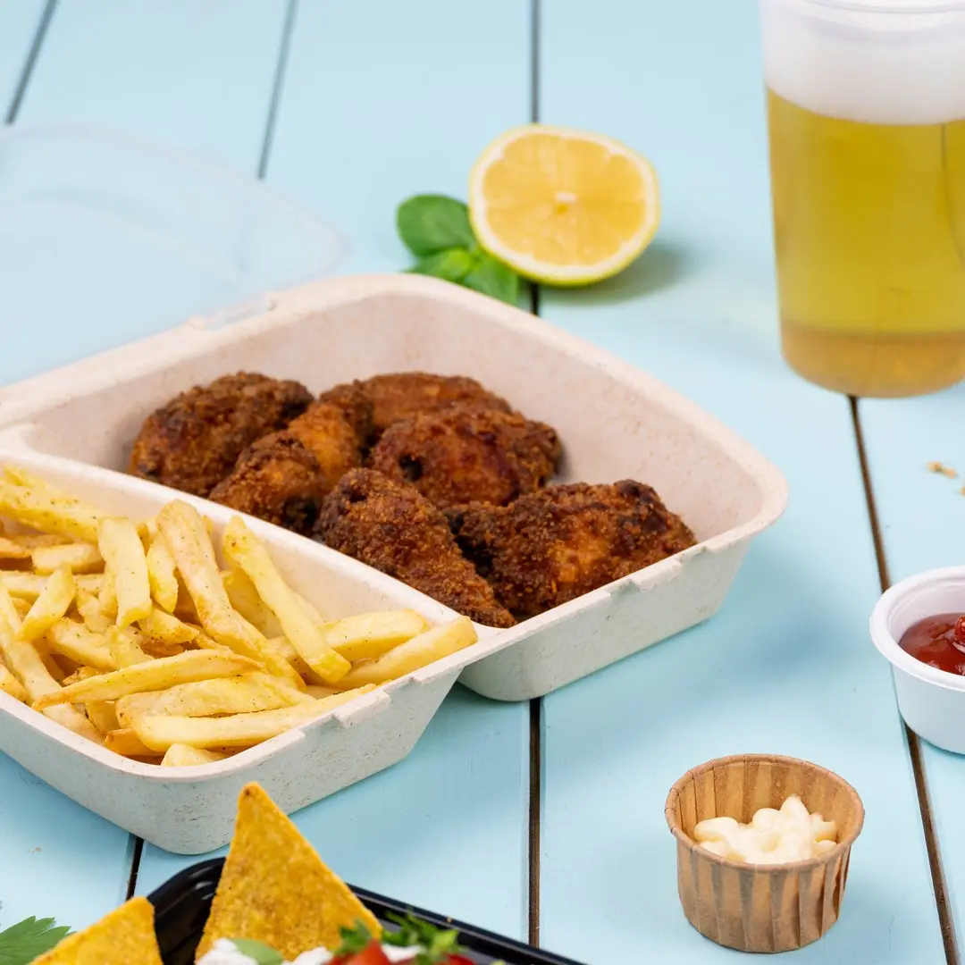 Takeout-Behälter mit gebratenem Hühnchen und Pommes, Zitronenscheibe, Bier und Dipsaucen auf einer blauen Holzoberfläche.