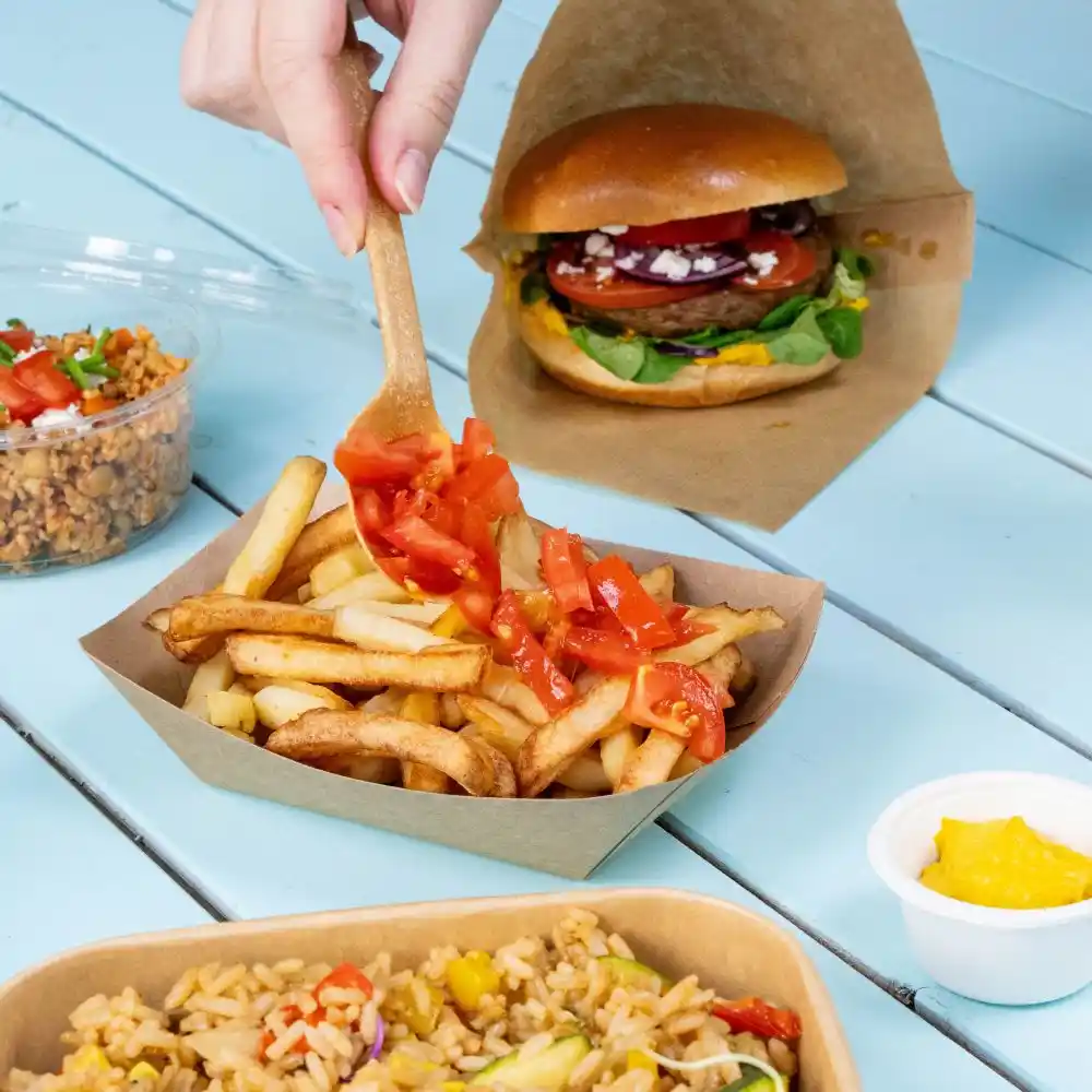 Eine Hand hält einen Holzlöffel und fügt gewürfelte Tomaten zu einer Portion Pommes auf einem braunen Tablett hinzu; im Hintergrund sind ein Burger und ein Becher Reis.