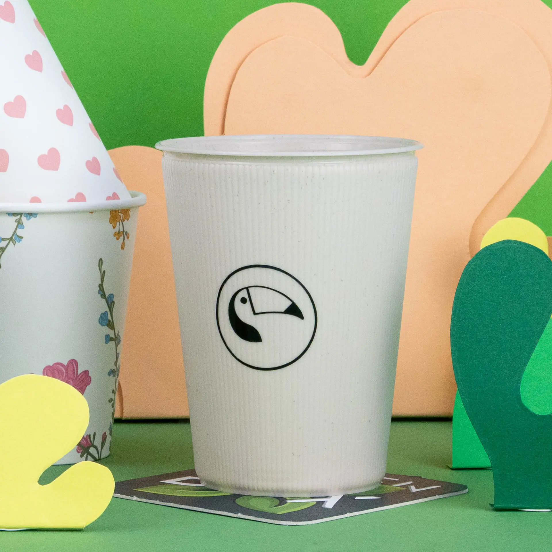 Weißer Wellen-Becher aus wiederverwendbarem Material mit einem Tukan-Logo, zwischen bunten Papierdekorationen auf grünem Hintergrund.