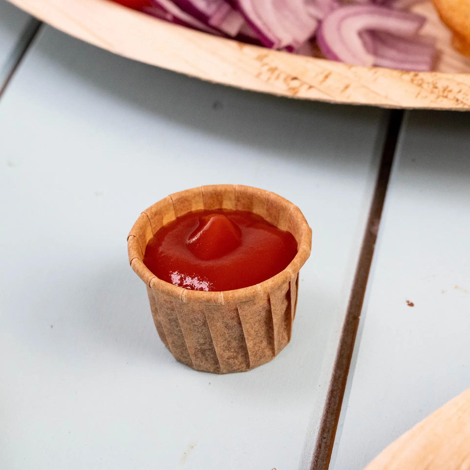 Brown paper cup filled with ketchup on a light blue surface, with red onions blurred in the background.