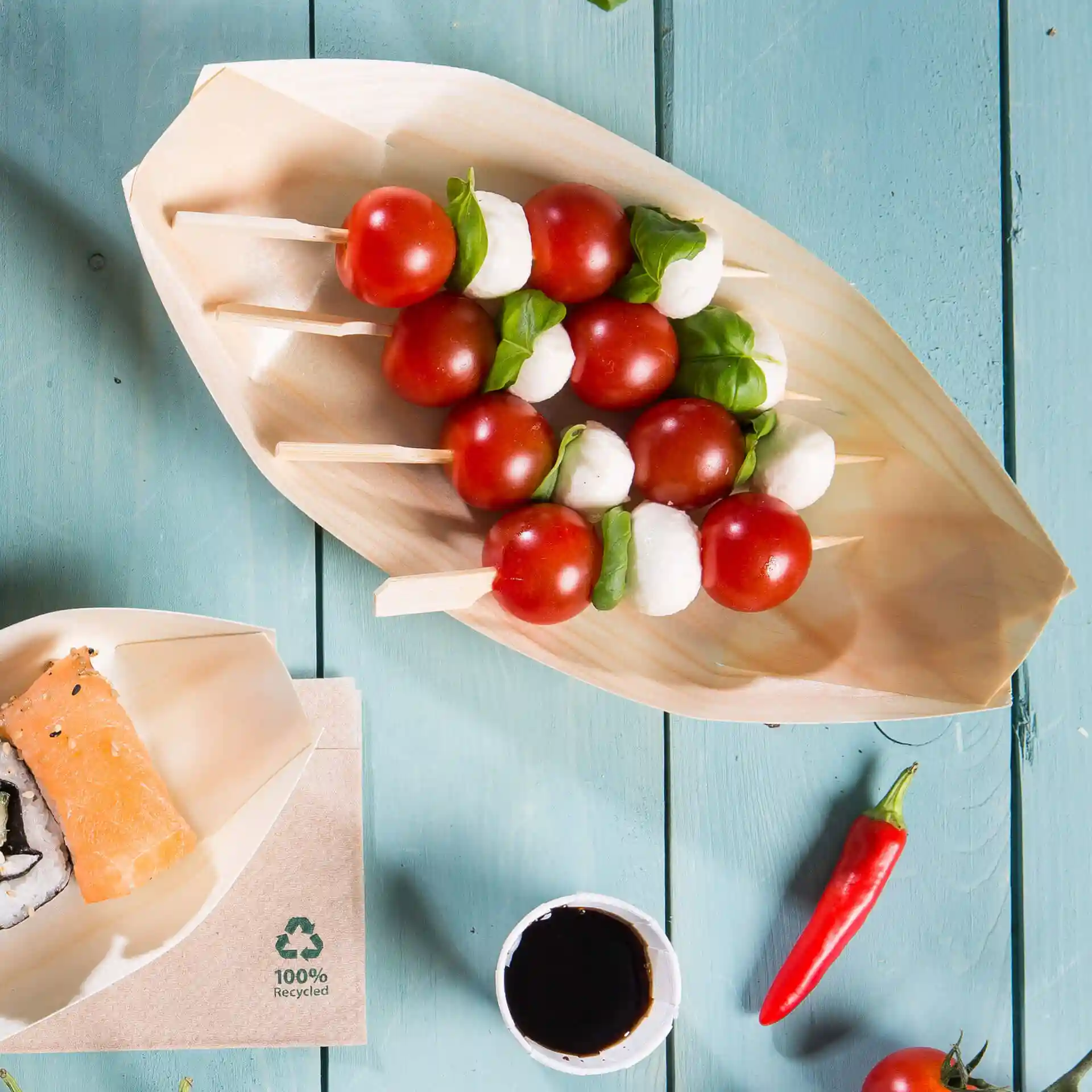 Wooden boat-shaped serving dish with skewers of cherry tomatoes, mozzarella, and basil, on a blue wooden surface.