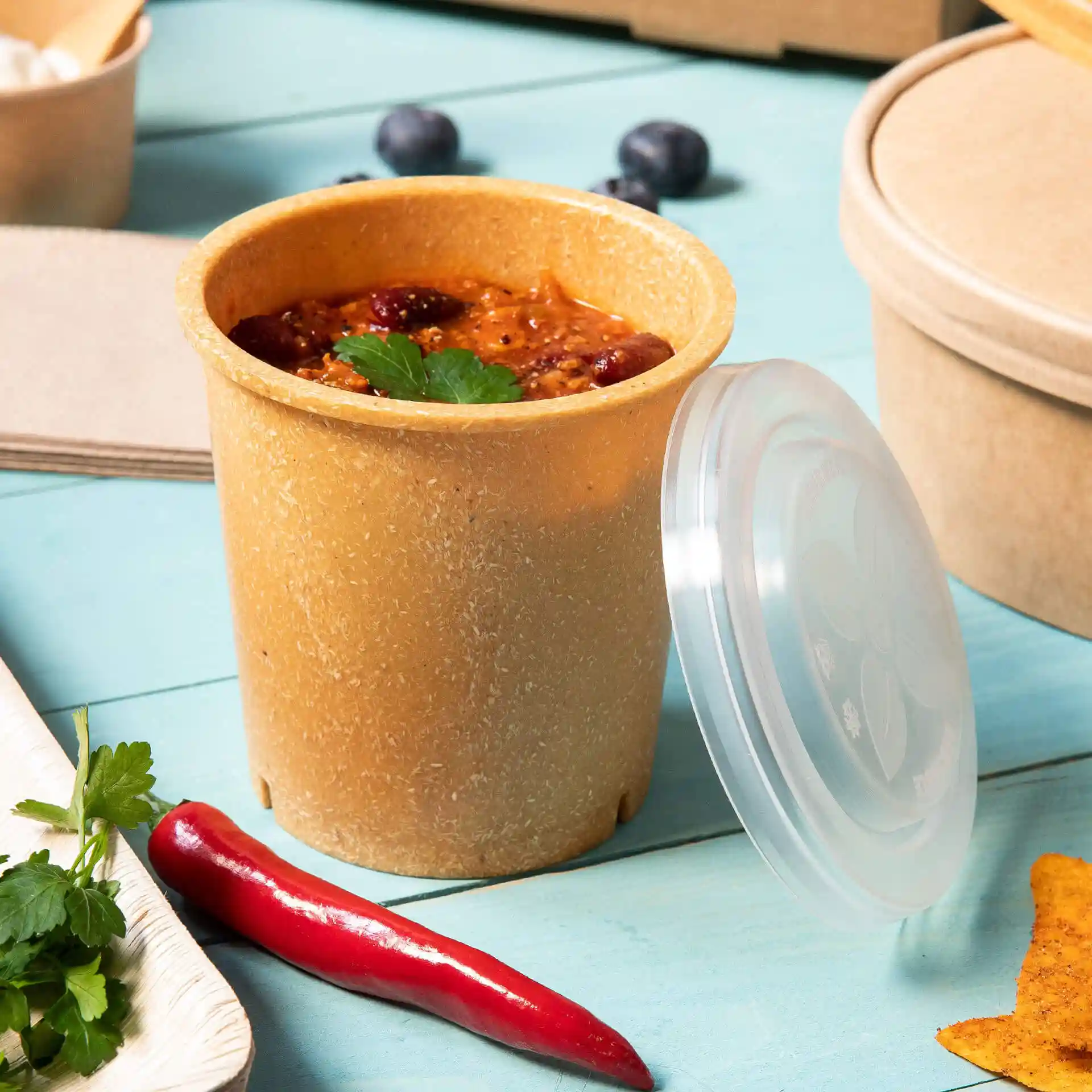 Brauner wiederverwendbarer Becher mit Deckel, gefüllt mit Essen, auf einer blauen Oberfläche mit einer roten Chili und Koriander daneben.