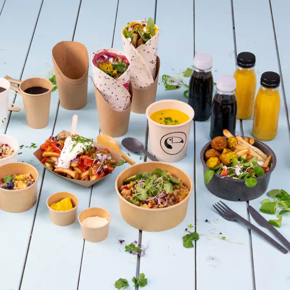 Verschiedene Lebensmittel in umweltfreundlicher Verpackung, darunter Wraps, Salate, Suppen und Getränke auf einer blauen Holzoberfläche.