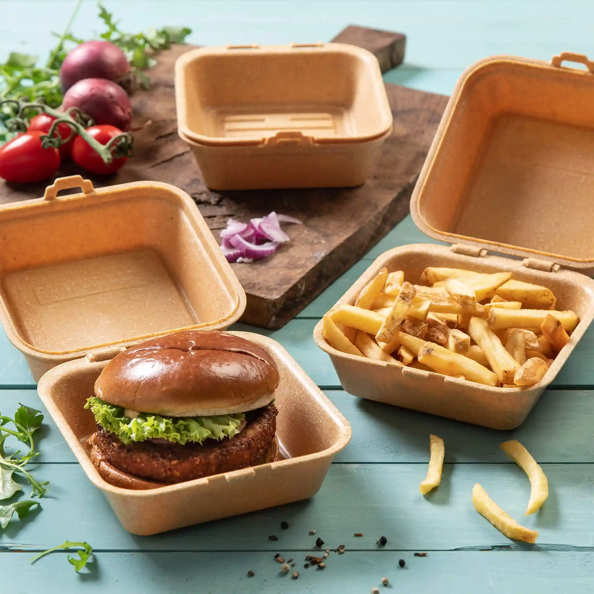 Braune wiederverwendbare Burgerbox mit Deckel, enthält einen Burger und Pommes, auf einer Holzoberfläche mit Gemüse daneben.