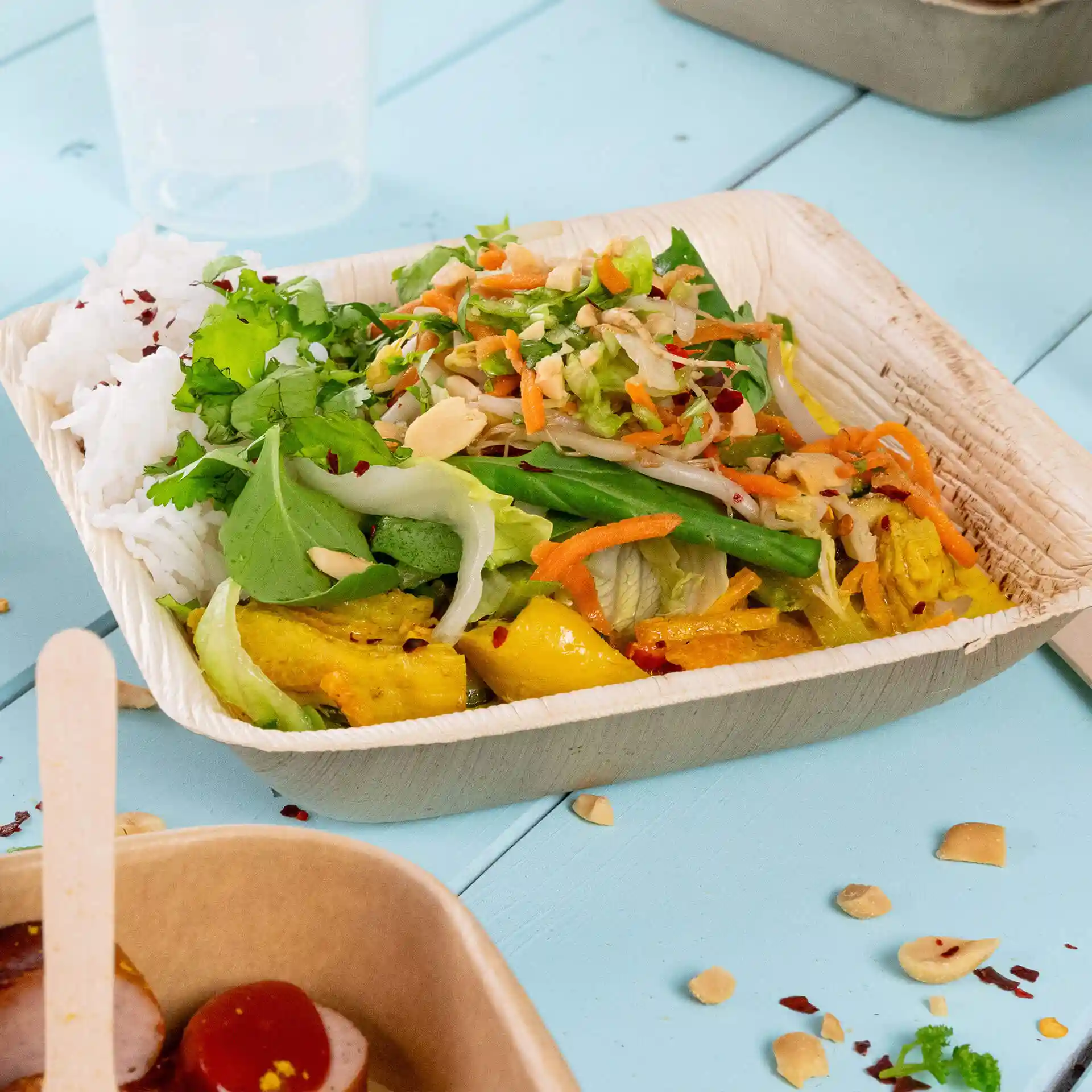 Square palm leaf bowl filled with rice, mixed salad, and vegetables on a blue surface.
