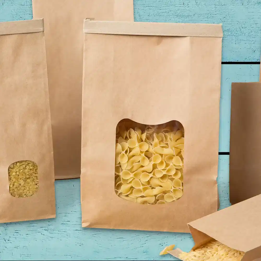Braune Papiertüten mit durchsichtigen Fenstern, gefüllt mit verschiedenen Pastasorten, auf einer blauen Holzoberfläche.