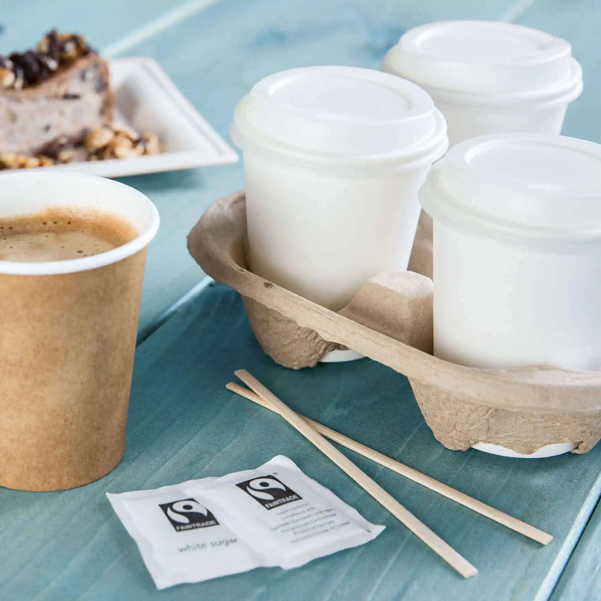 Image of a brown paper cup with coffee, three white cups in a holder, wooden stirrers, and sugar packets on a blue surface.