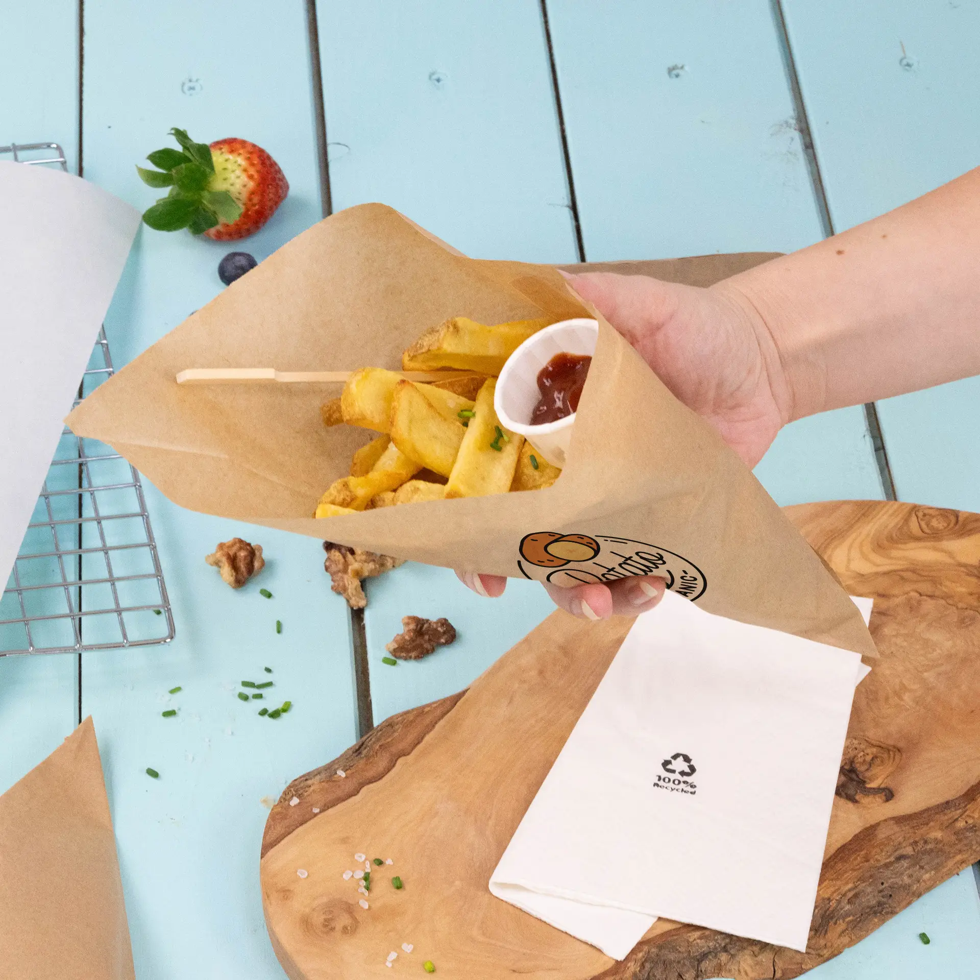 Hand hält einen braunen Papierbecher mit Pommes frites und einem kleinen Becher Ketchup, auf einer blauen Holzoberfläche.