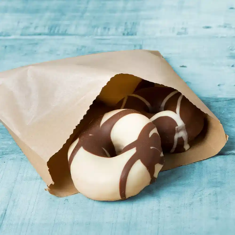 Schokoladenüberzogene Donuts, teilweise sichtbar in einer braunen Papiertüte auf einer blauen Holzoberfläche.
