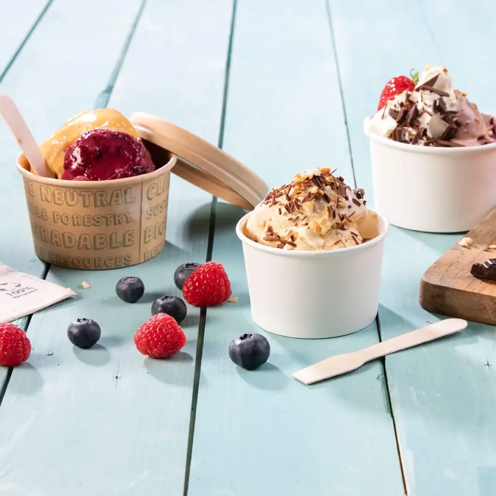 Ice cream cups with different flavors, fresh raspberries, blueberries, and a wooden spatula on a blue wooden surface.